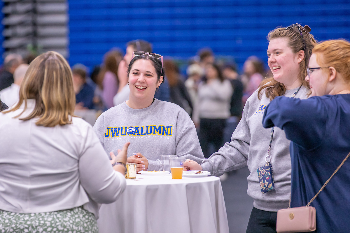 Three individuals engage in lively conversation around a table at an event, with one wearing a "JWU ALUMNI" sweatshirt and another smiling broadly while holding a drink.