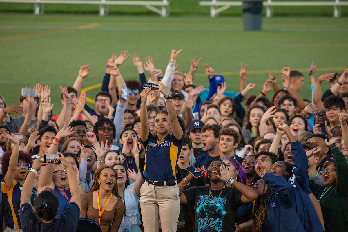 A crowd of young people at a sports event cheer enthusiastically, raising their hands and phones in excitement.