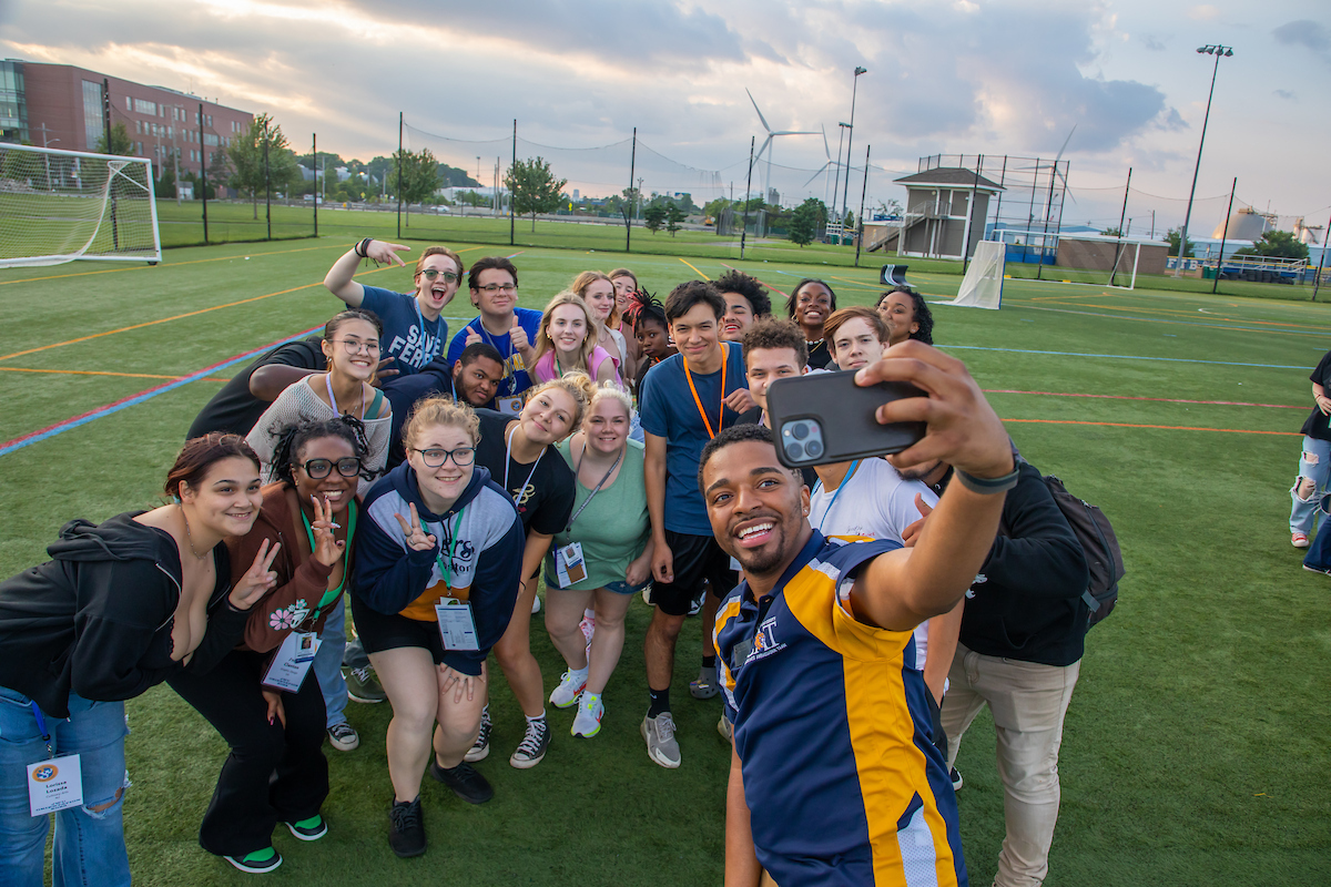 A diverse group of young people pose together on a sports field, taking a selfie with one person holding a smartphone.