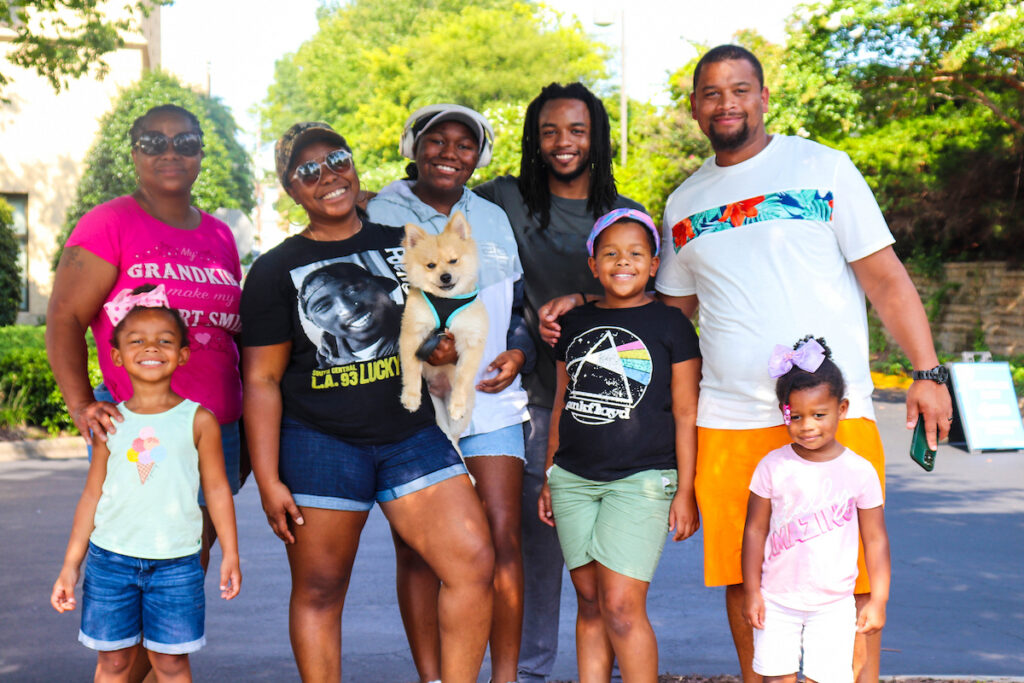 A group of adults and children, including a small dog, pose together outdoors on a sunny day.