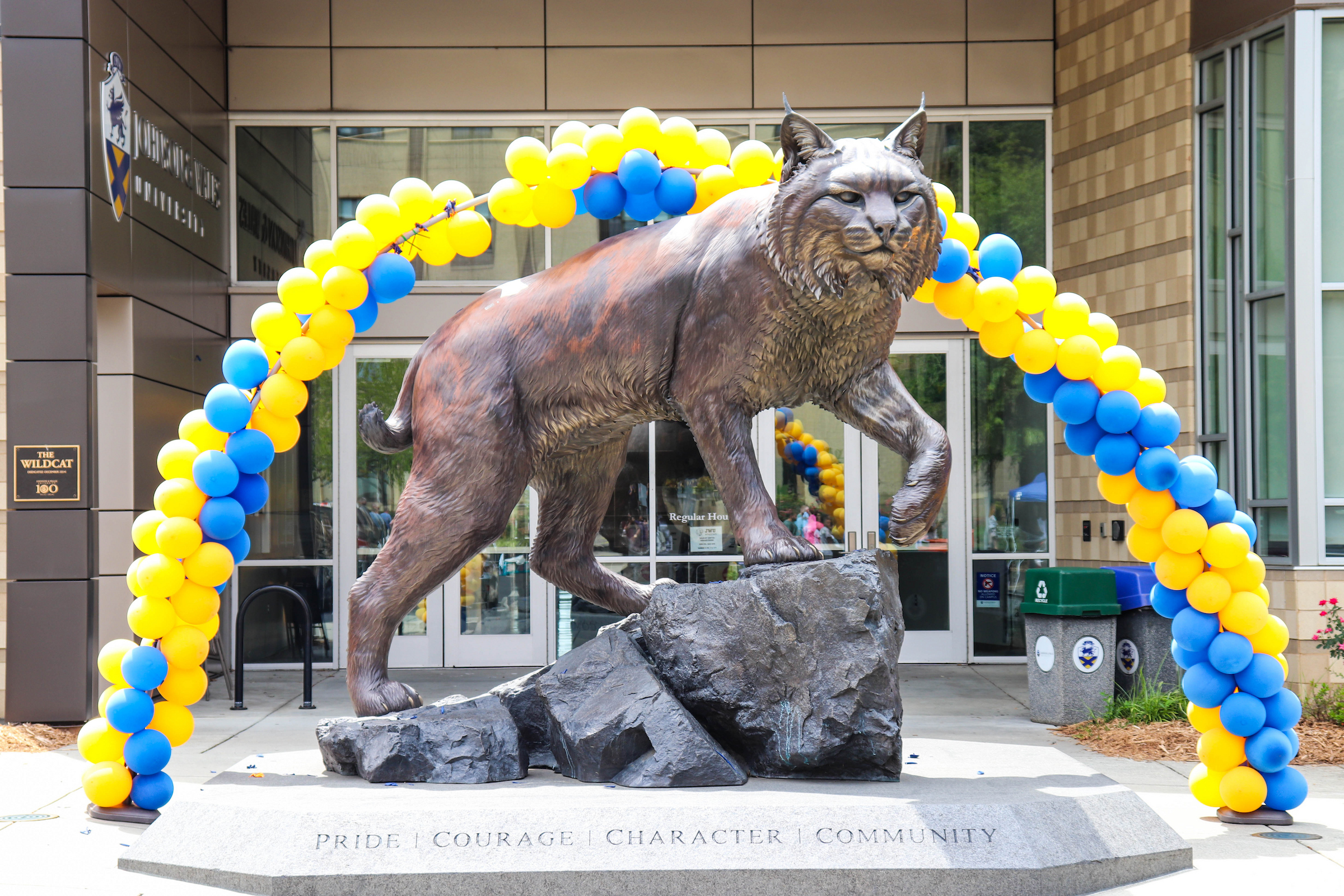 A bronze statue of a bobcat stands on a rocky base beneath a blue and yellow balloon arch, with the words "PRIDE | COURAGE | CHARACTER | COMMUNITY" inscribed on the pedestal.