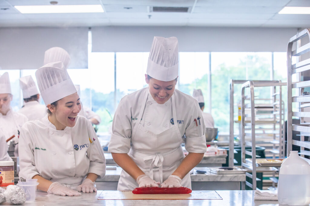 Two culinary students in a professional kitchen laugh while working together to shape red fondant.