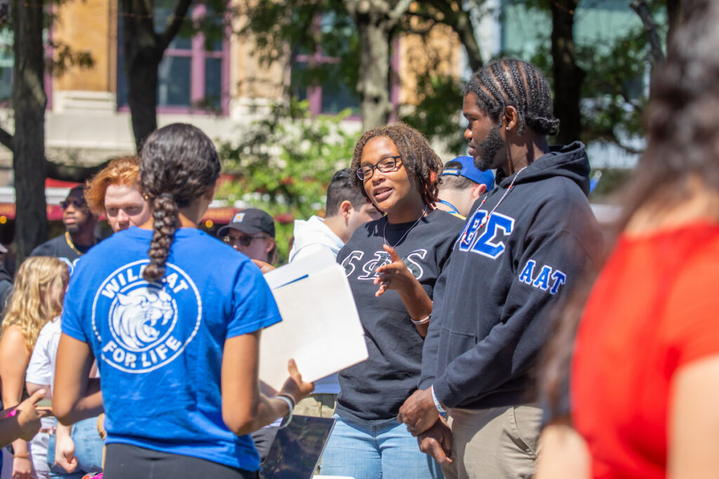 A group of people gather outdoors, with one person in a blue shirt speaking to others, including someone in a dark hoodie with Greek letters.
