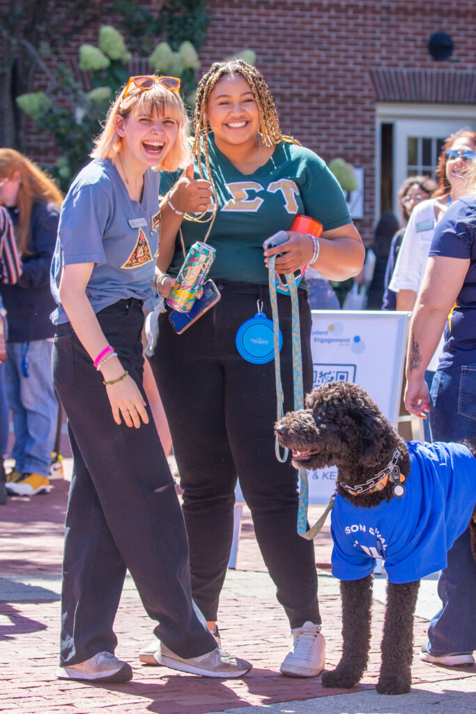 Two smiling individuals pose with a curly-haired dog wearing a blue shirt, holding leashes and accessories, at an outdoor event.