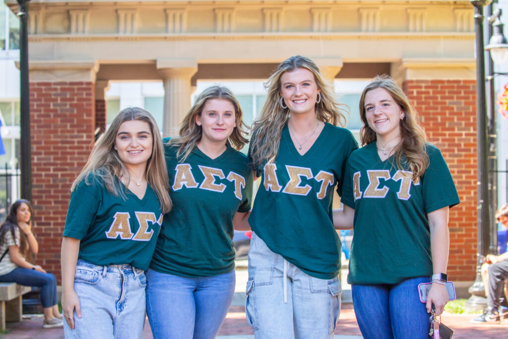 Four individuals wearing green shirts with Greek letters stand together outdoors in front of a brick building.