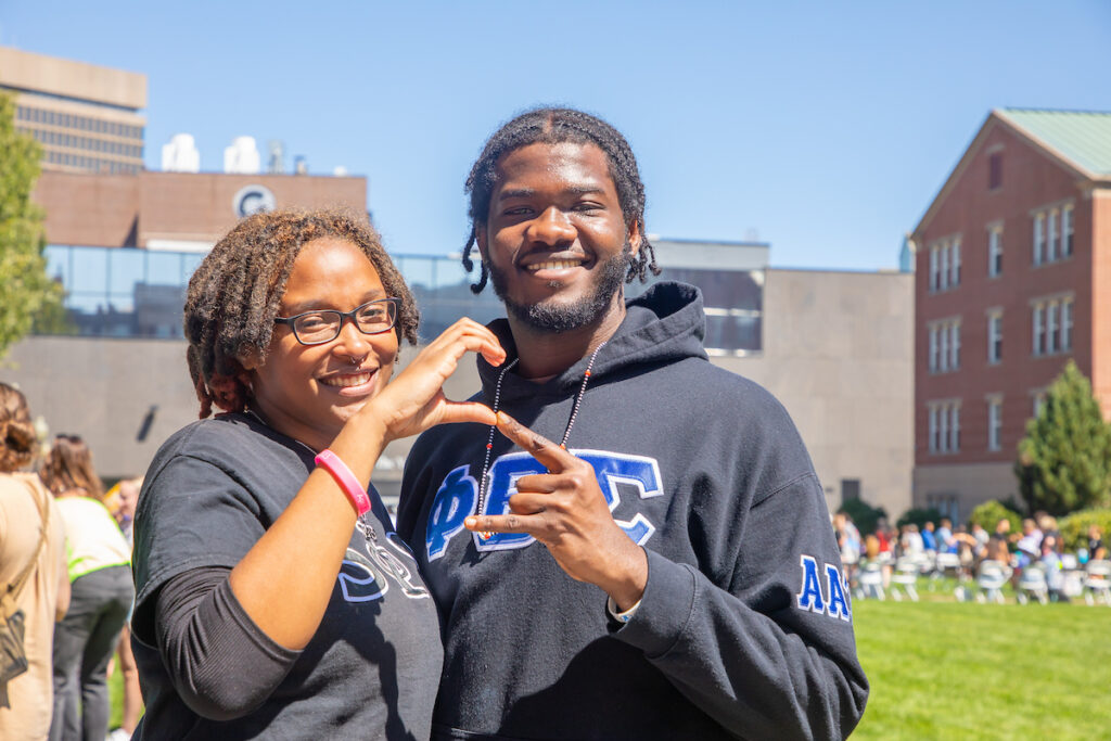 Two individuals in matching hoodies make a heart shape with their hands while smiling outdoors on a sunny day.