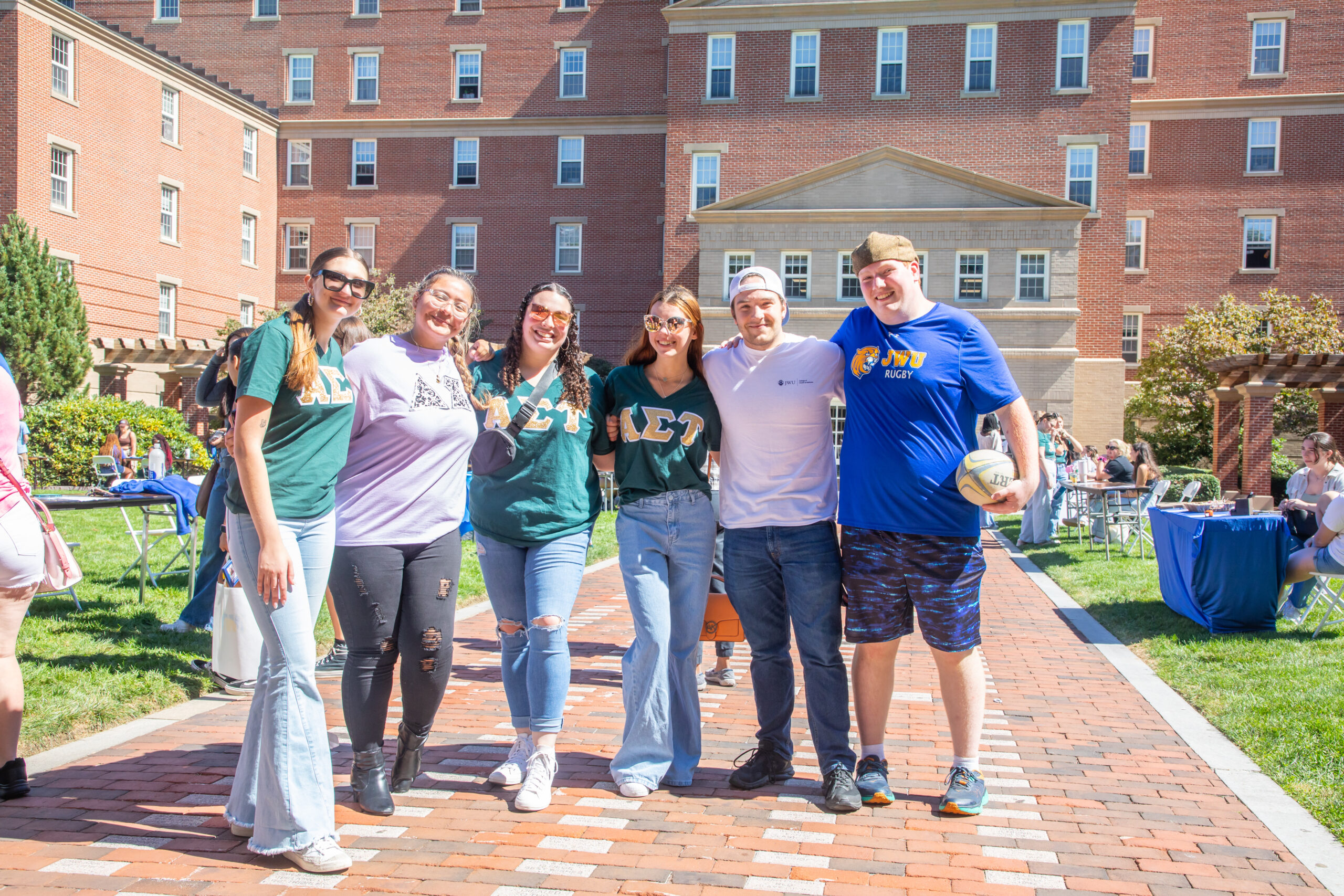 A group of six people pose together on a brick walkway in front of a large brick building, with one person holding a rugby ball.