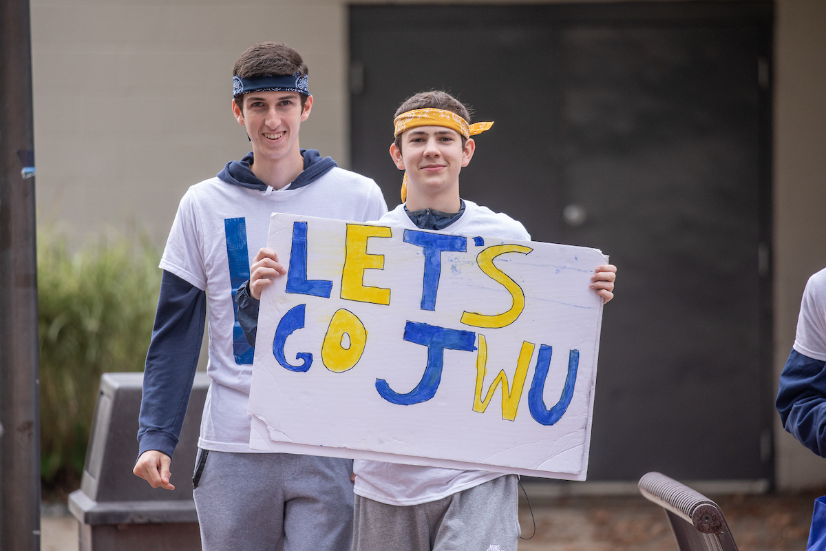 Two individuals wearing headbands hold a handmade sign that reads "LET'S GO JWU" in blue and yellow letters.