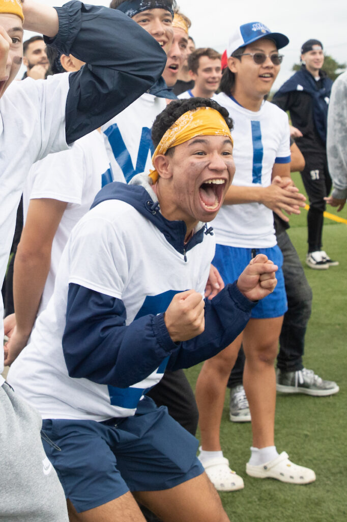 A group of young people in athletic attire celebrate enthusiastically on a grass field, with one person in the foreground cheering with fists raised.