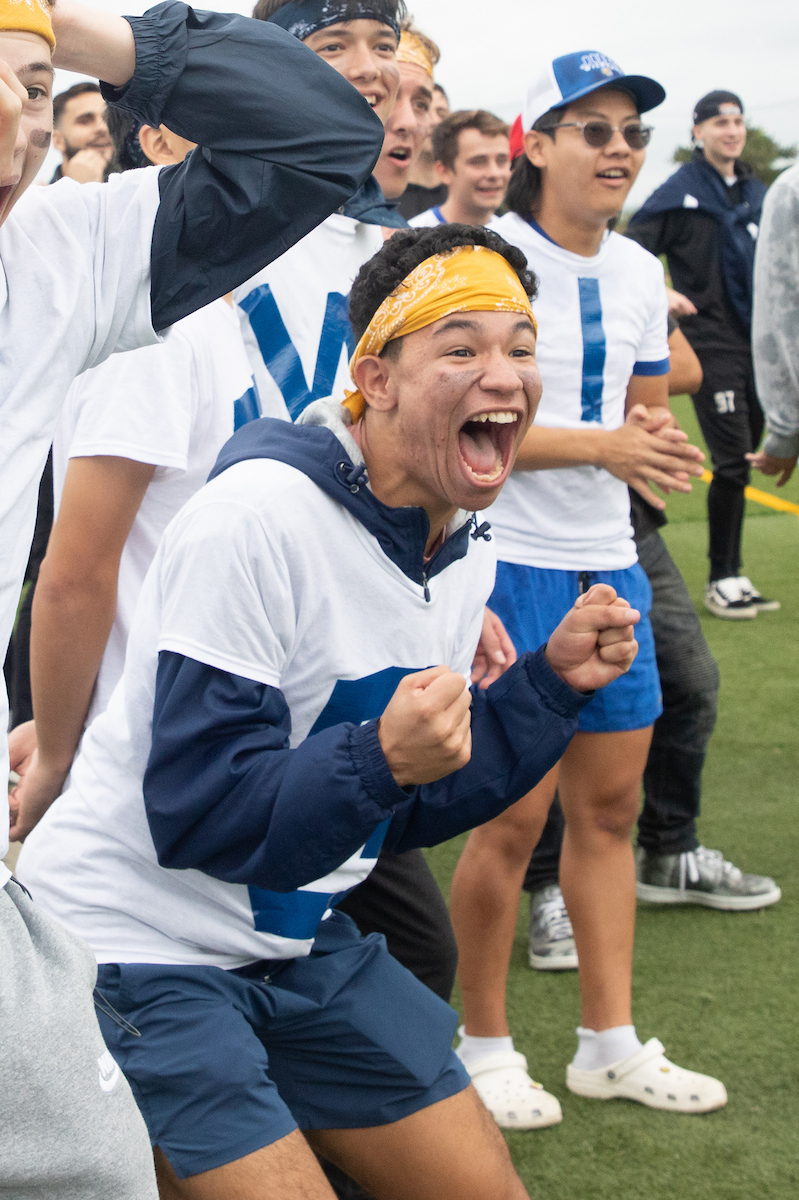 A group of young people in athletic attire celebrate enthusiastically on a grass field, with one person in the foreground cheering with fists raised.
