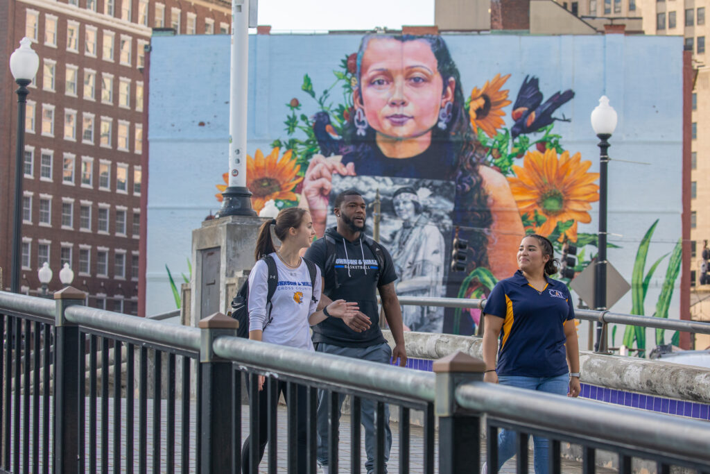 Three people walk across a bridge, passing by a large mural depicting a young person surrounded by sunflowers and birds.