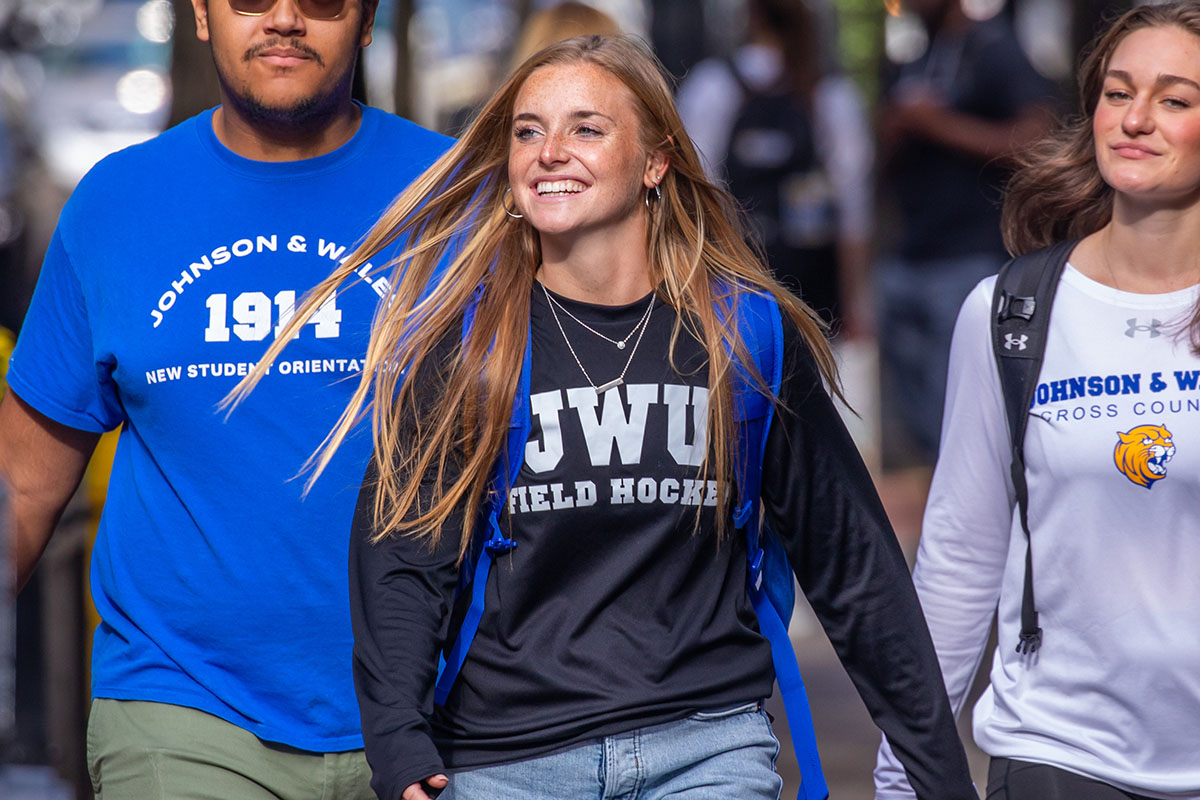 Three individuals wearing Johnson & Wales University apparel walk together outdoors, with the central person smiling broadly.