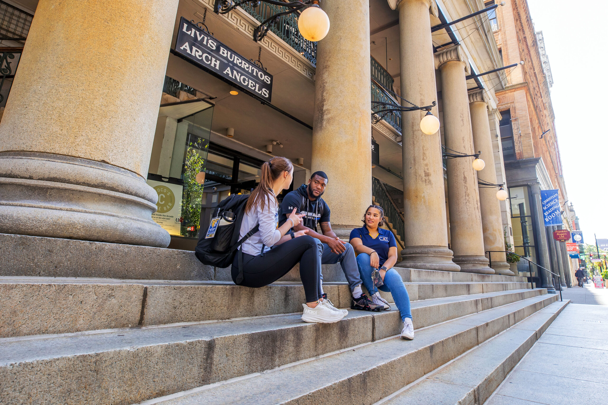 Three individuals sit on stone steps outside a building with large columns, engaged in conversation under bright daylight.
