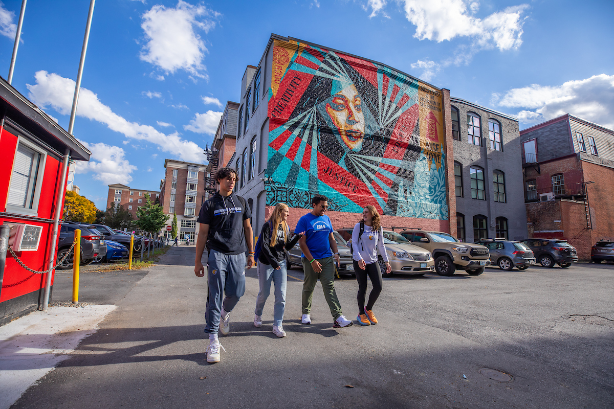 Four individuals stroll across a paved area under a bright blue sky, passing a large, colorful mural on a brick building.
