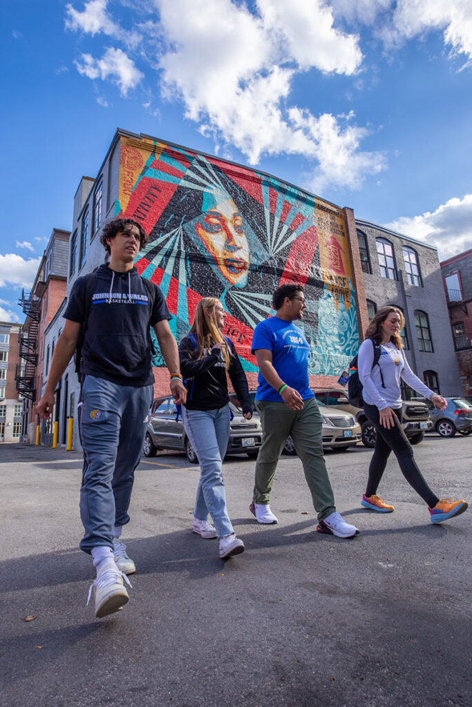 Four individuals walk across a street under a bright blue sky, passing a large colorful mural on a brick building.