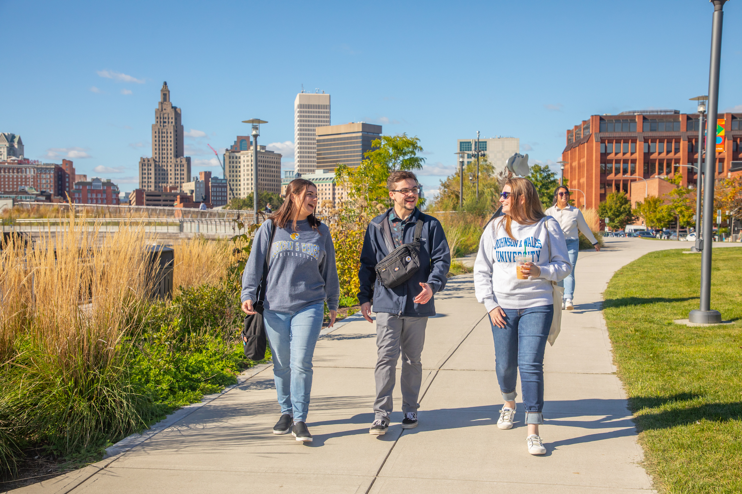 Three individuals stroll along a paved path lined with grass and tall plants, with a city skyline visible in the background under a clear blue sky.