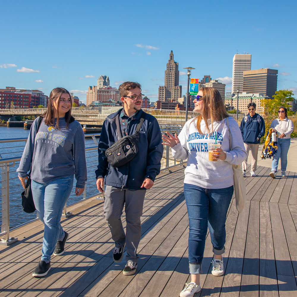 Three students walk along a wooden boardwalk beside a river, with the Providence skyline visible in the background under a clear blue sky.
