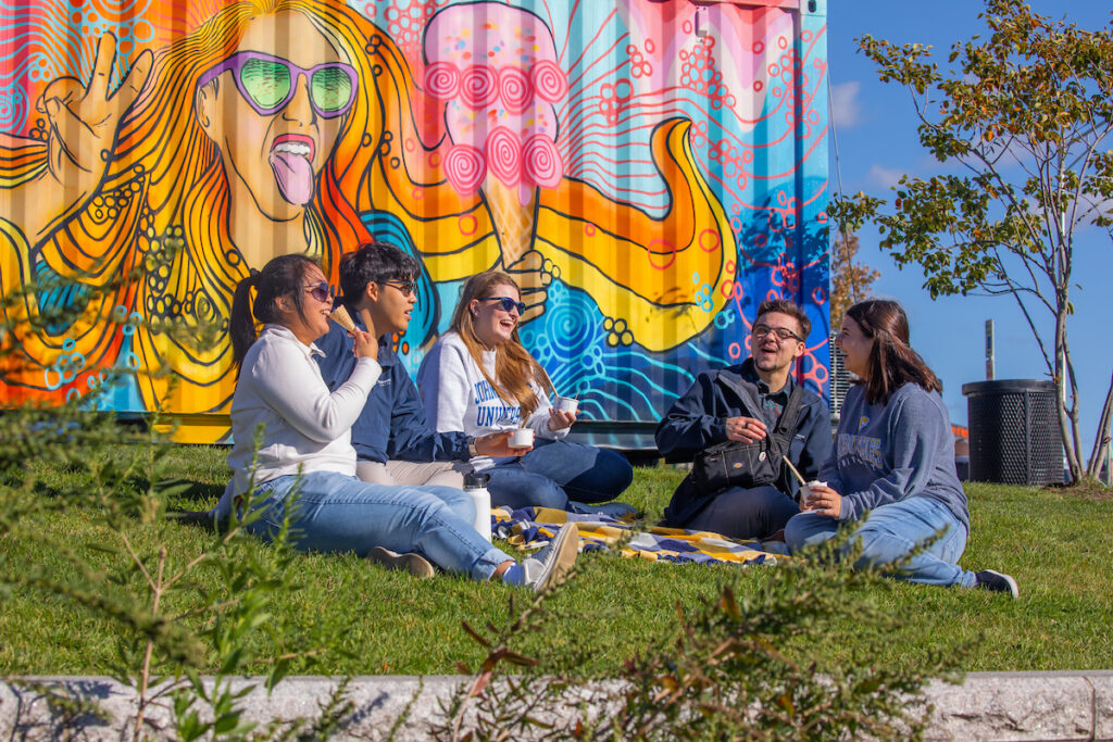 A group of five individuals relaxes on a grassy area, enjoying snacks and conversation in front of a vibrant, colorful mural on a shipping container.