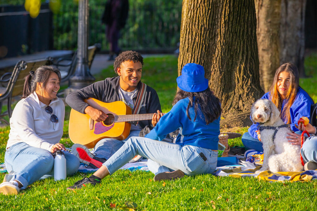 A group of friends relaxing on a blanket in a park, listening to one student playing the guitar.