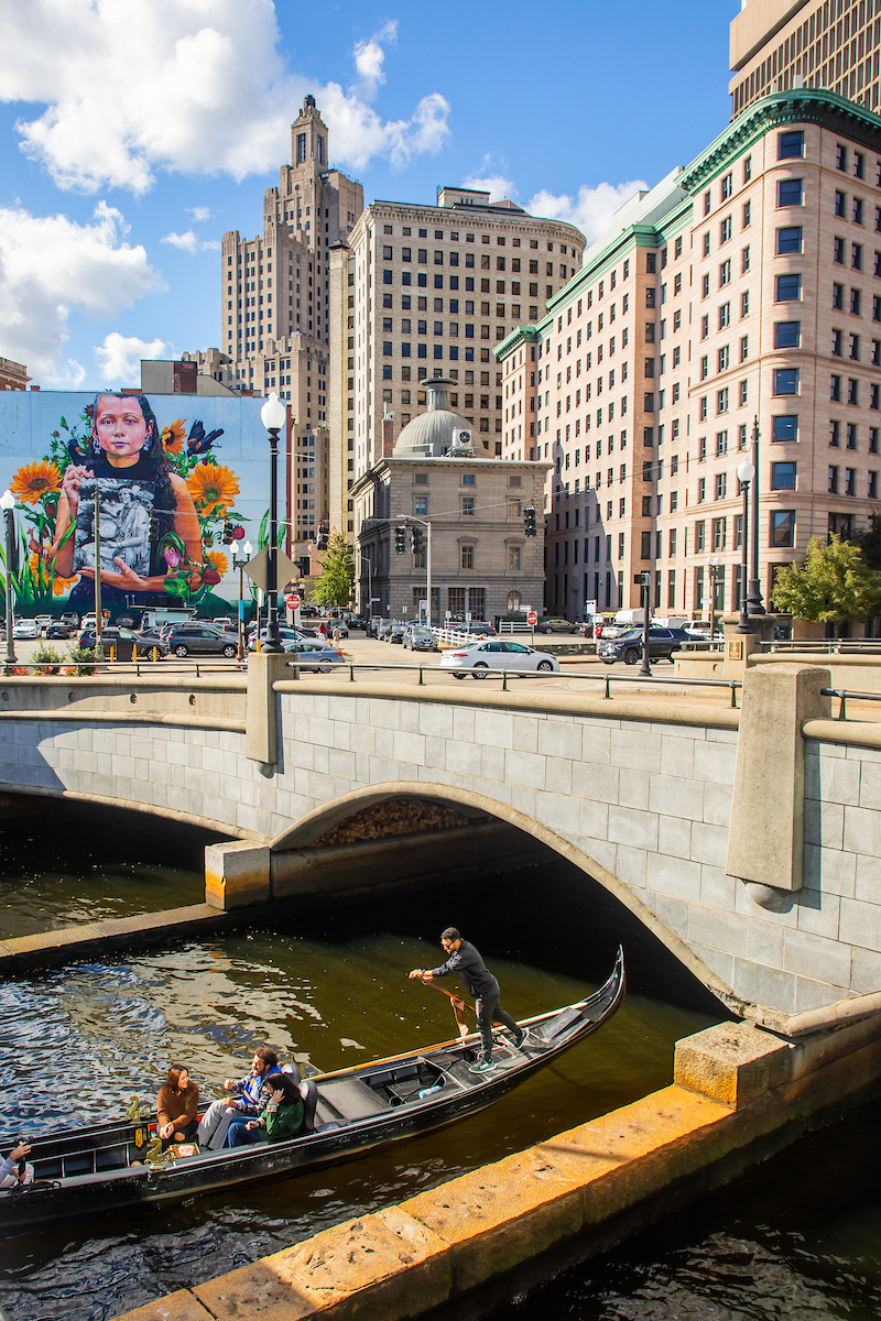 A gondolier navigates a narrow canal beneath a stone bridge, passing by a large mural and tall city buildings under a bright blue sky.