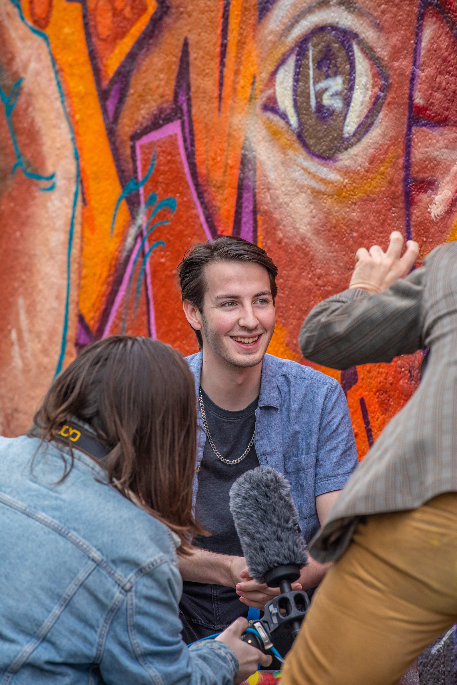 A person smiles while being interviewed outdoors, with a vibrant graffiti mural featuring a large eye in the background.