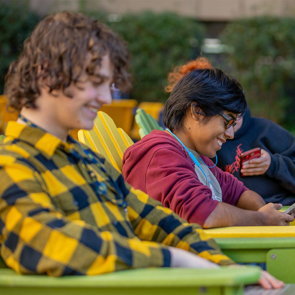 Two students relax in colorful outdoor chairs, smiling and engaged with their phones.