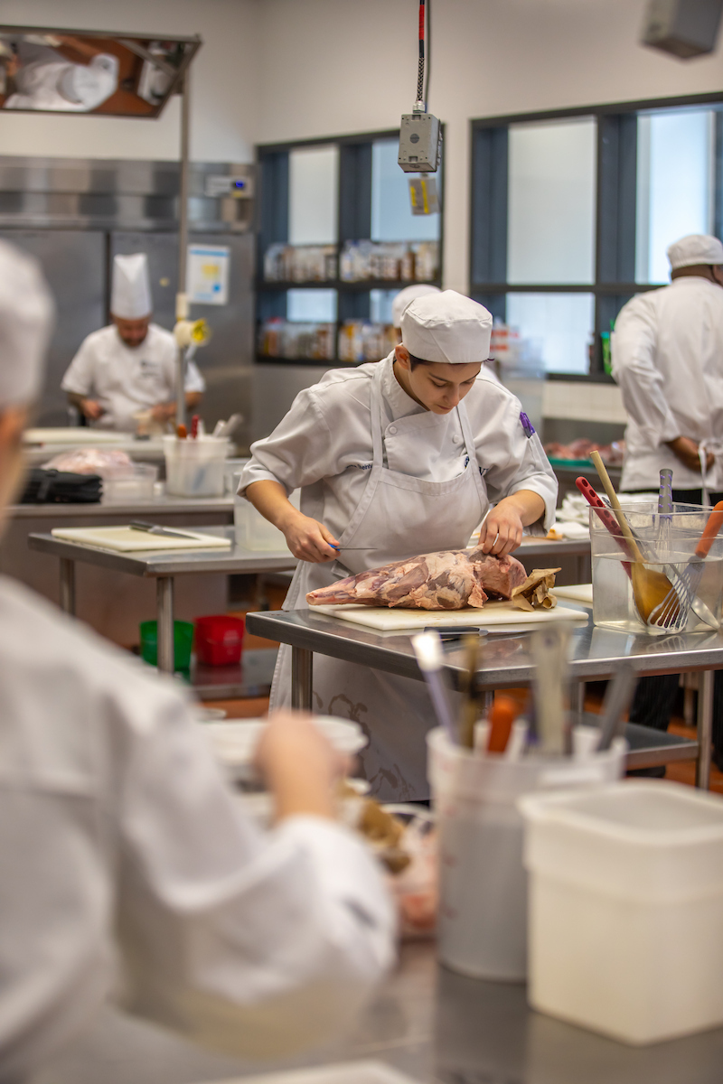 A chef in a professional kitchen carefully trims a large piece of meat on a cutting board while other staff members work in the background.