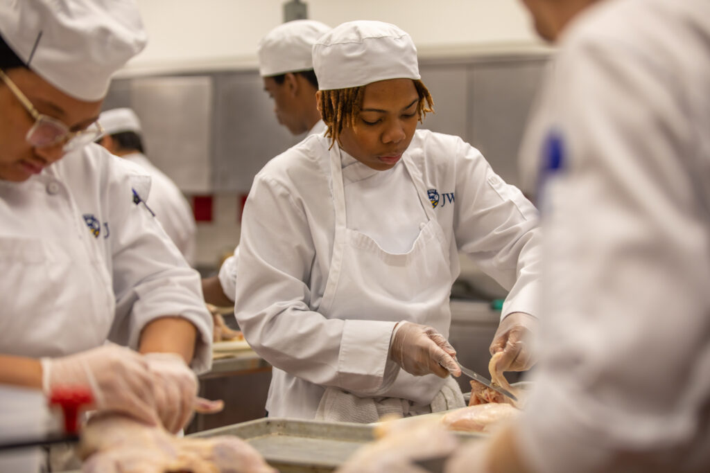Several individuals in white chef uniforms and hats are preparing meat in a kitchen.