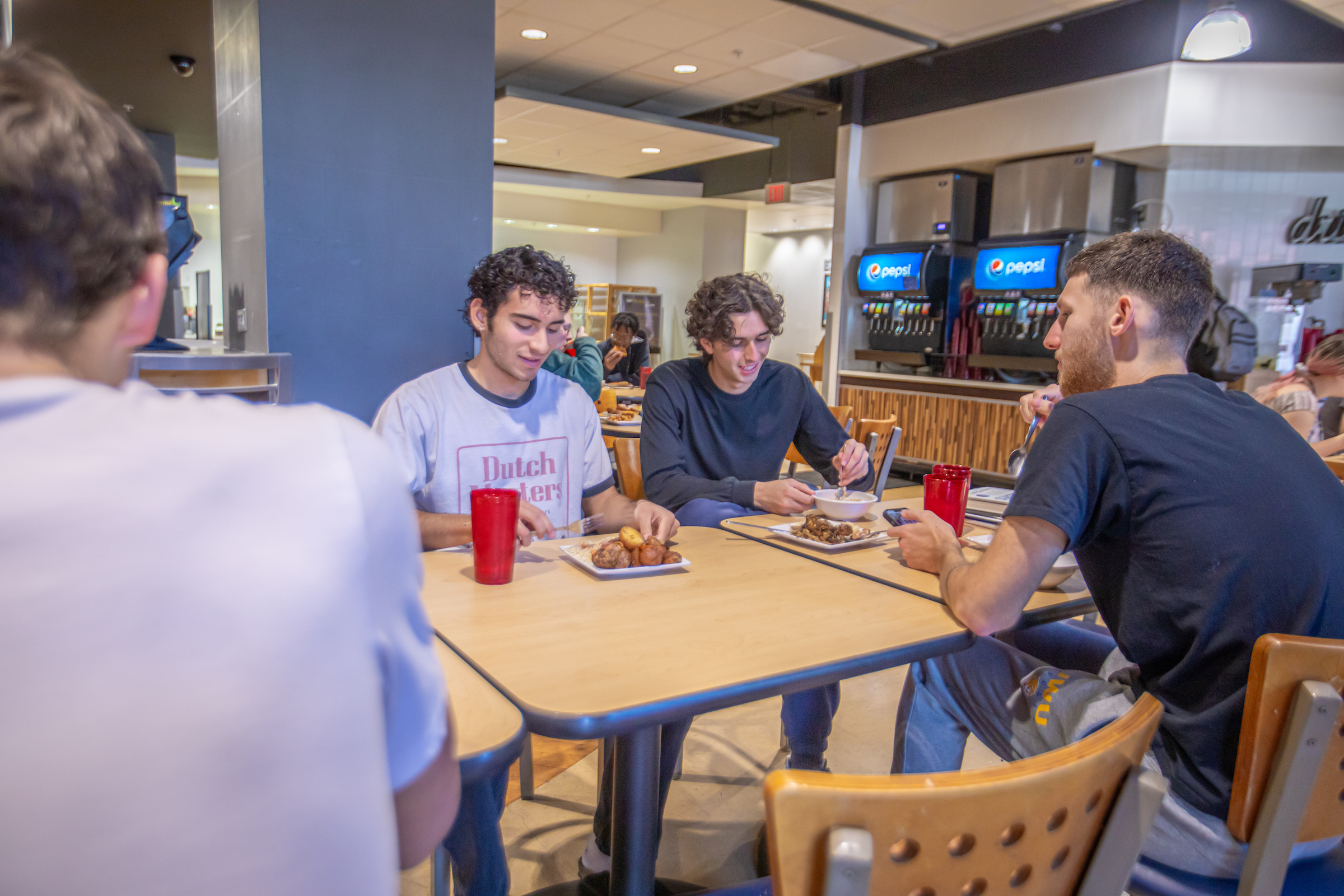 Four individuals are seated at a table in a cafeteria, eating and conversing while surrounded by food and drink options.