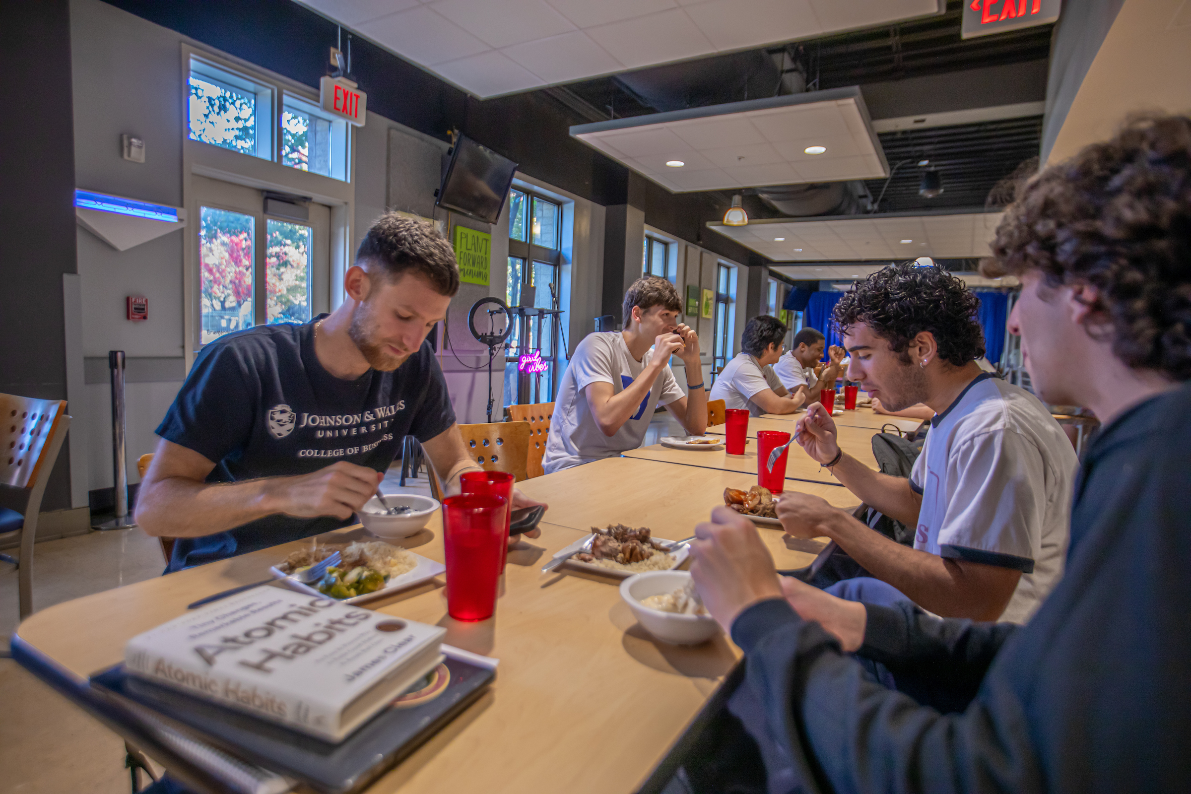 Several individuals are seated at a long table in a modern dining area, eating meals and engaging in conversation.