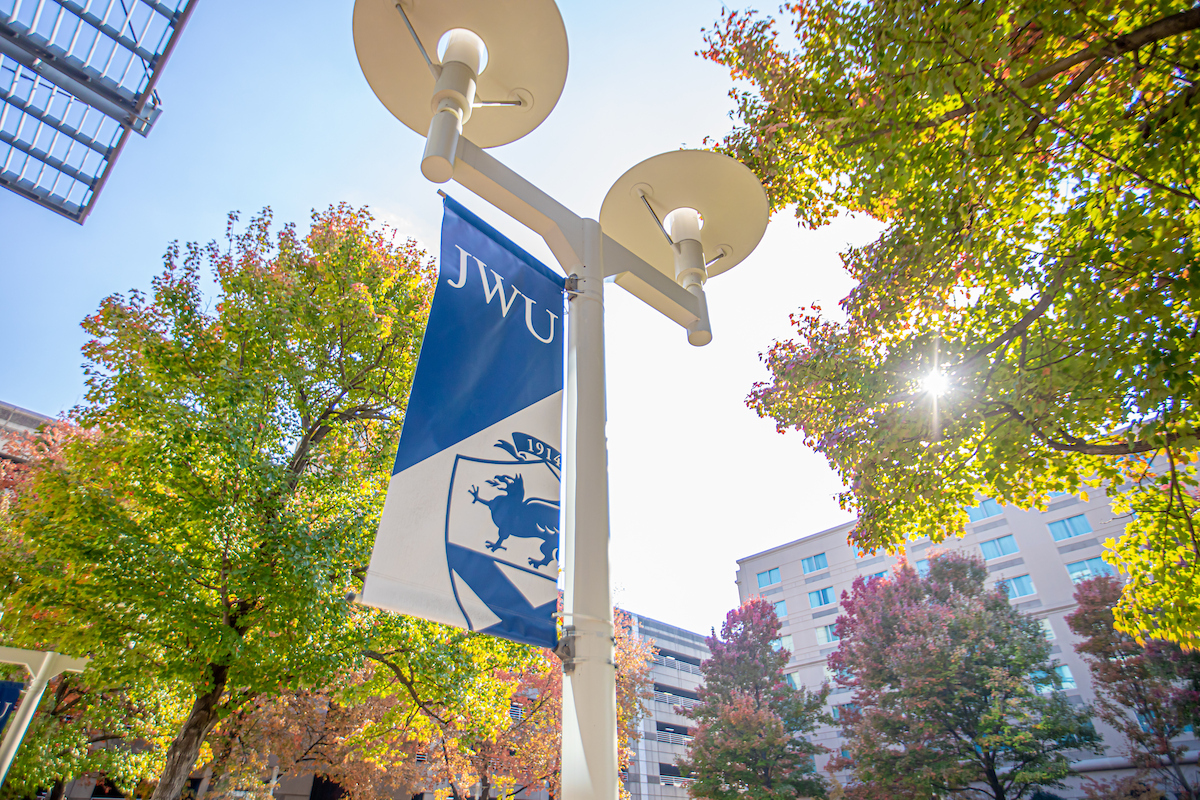 A blue and white banner with "JWU" and a crest hangs from a modern streetlight against a backdrop of autumn trees and a bright sky.