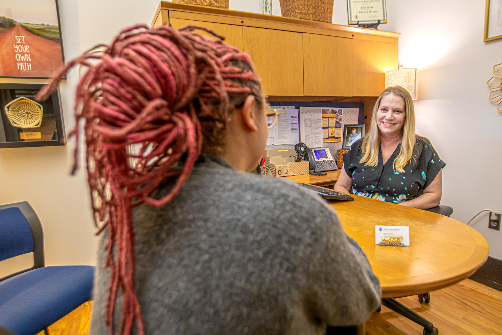 A counselor speaks with a student across a wooden desk in a warmly lit office setting.