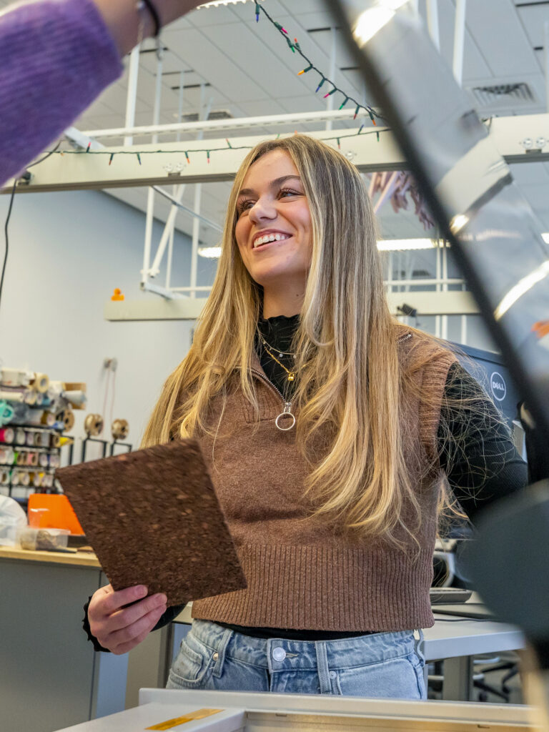 A person with long blonde hair smiles while holding a brown textured panel in a workshop setting.