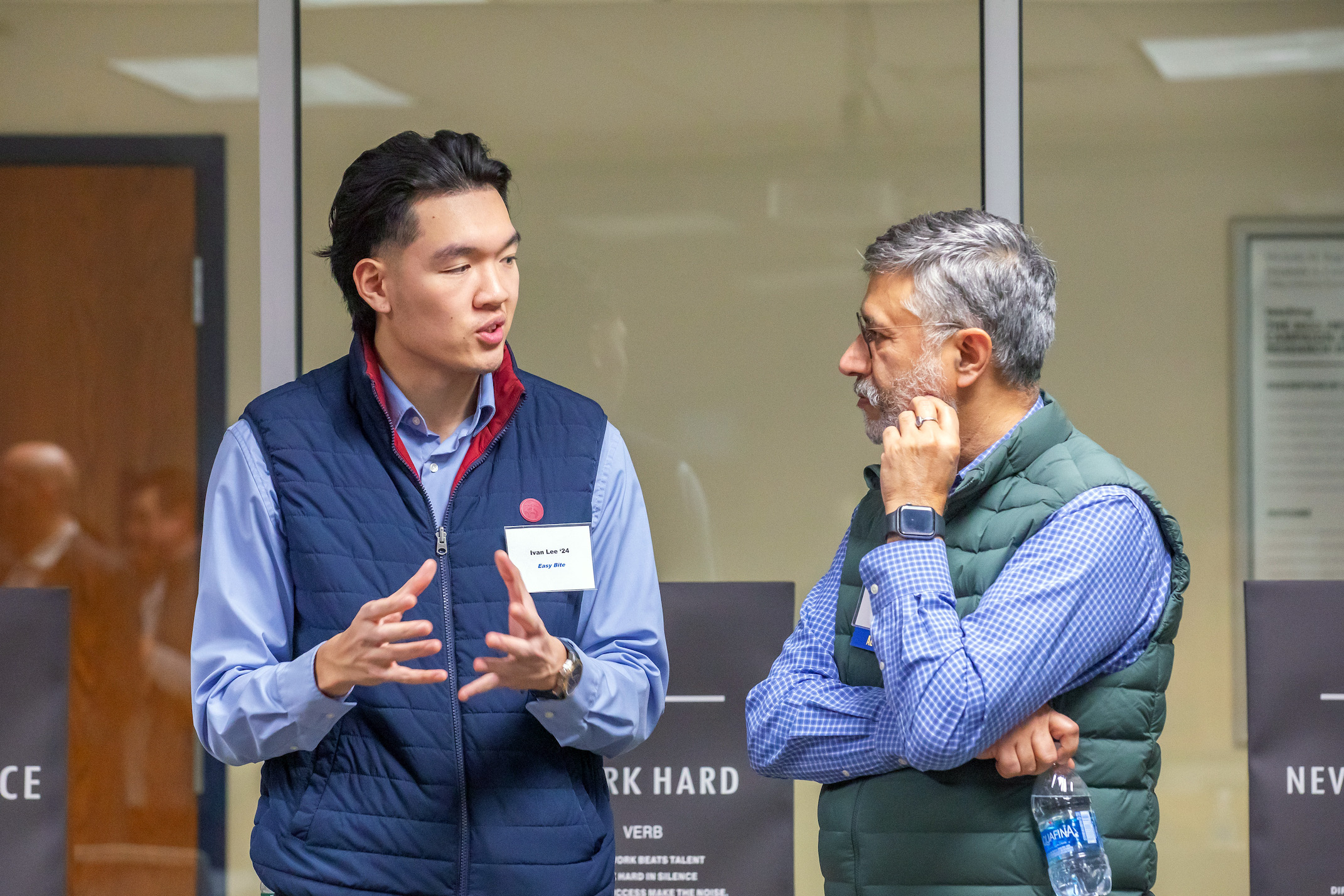 Two individuals engage in conversation indoors, one gesturing while speaking and the other listening thoughtfully with a water bottle in hand.