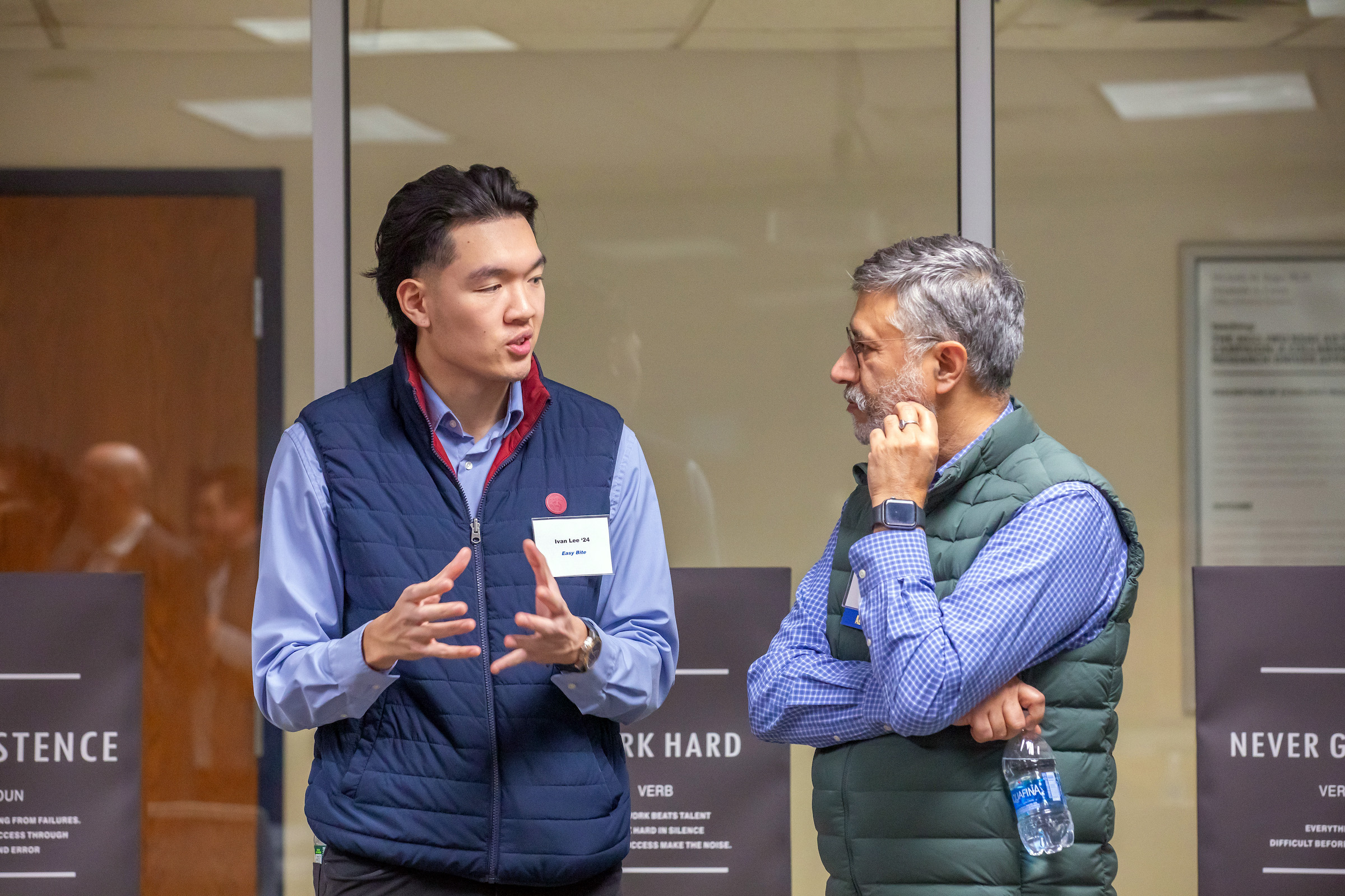 Two individuals engage in conversation indoors, one gesturing while speaking and the other listening thoughtfully with a water bottle in hand.