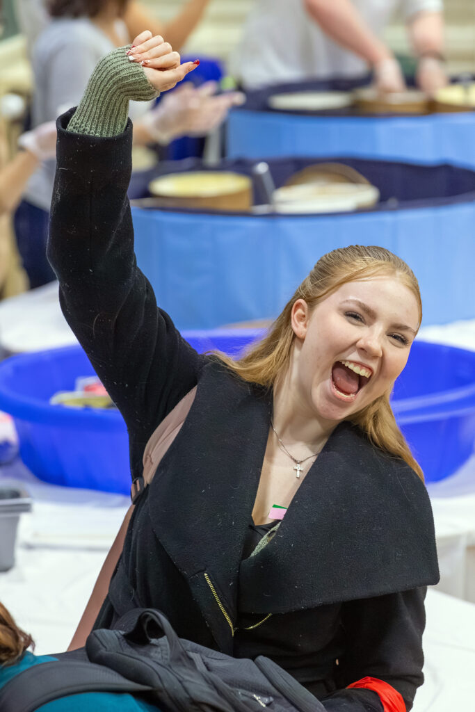 A person with long hair, wearing a black coat and green knit glove, raises their fist in excitement while laughing joyfully at an event.