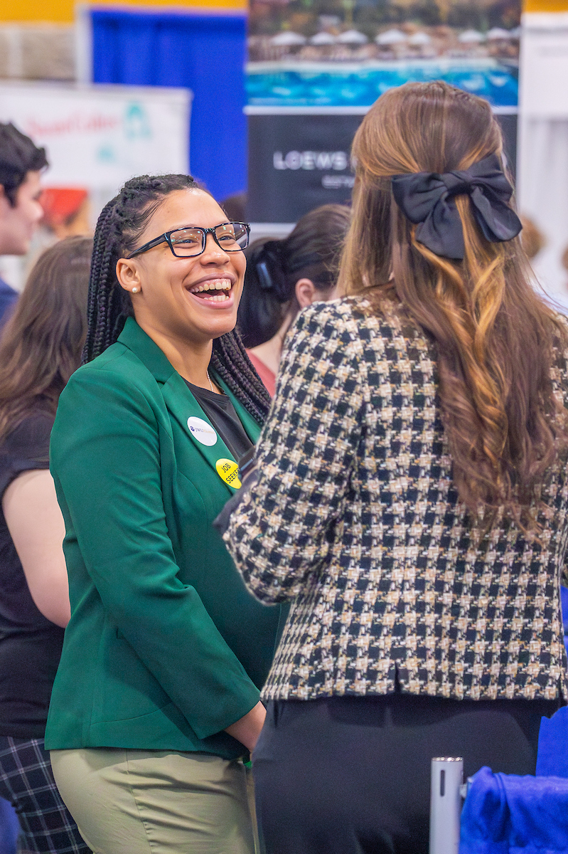 A person in a green blazer laughs while conversing with another individual at what appears to be a job fair or professional event.