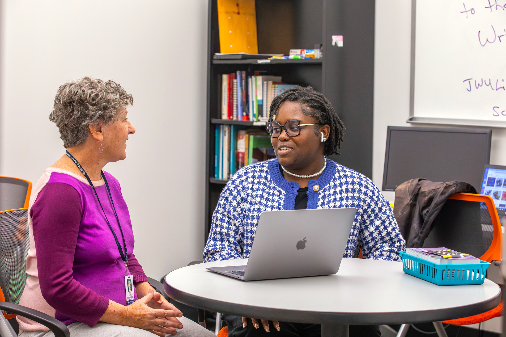 Two individuals engage in conversation at a table with a laptop, in a room with bookshelves and a whiteboard.