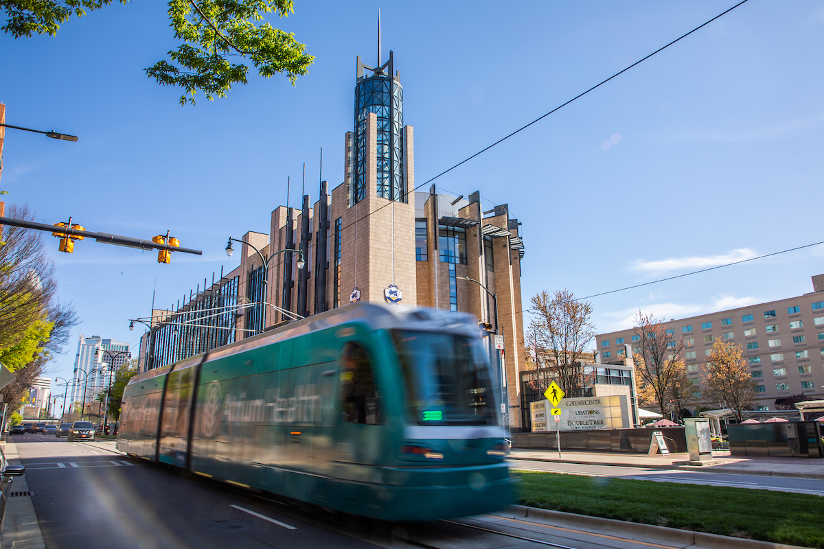 A teal streetcar speeds past a modernist building with a tall spire under a clear blue sky.