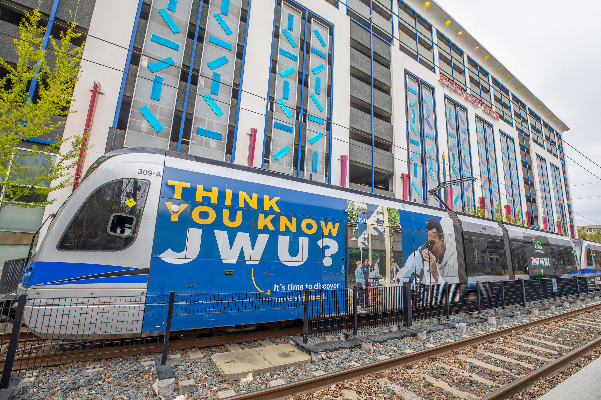 A modern light rail train with a blue advertisement for JWU passes by a multi-story building with distinctive blue and gray architectural features.