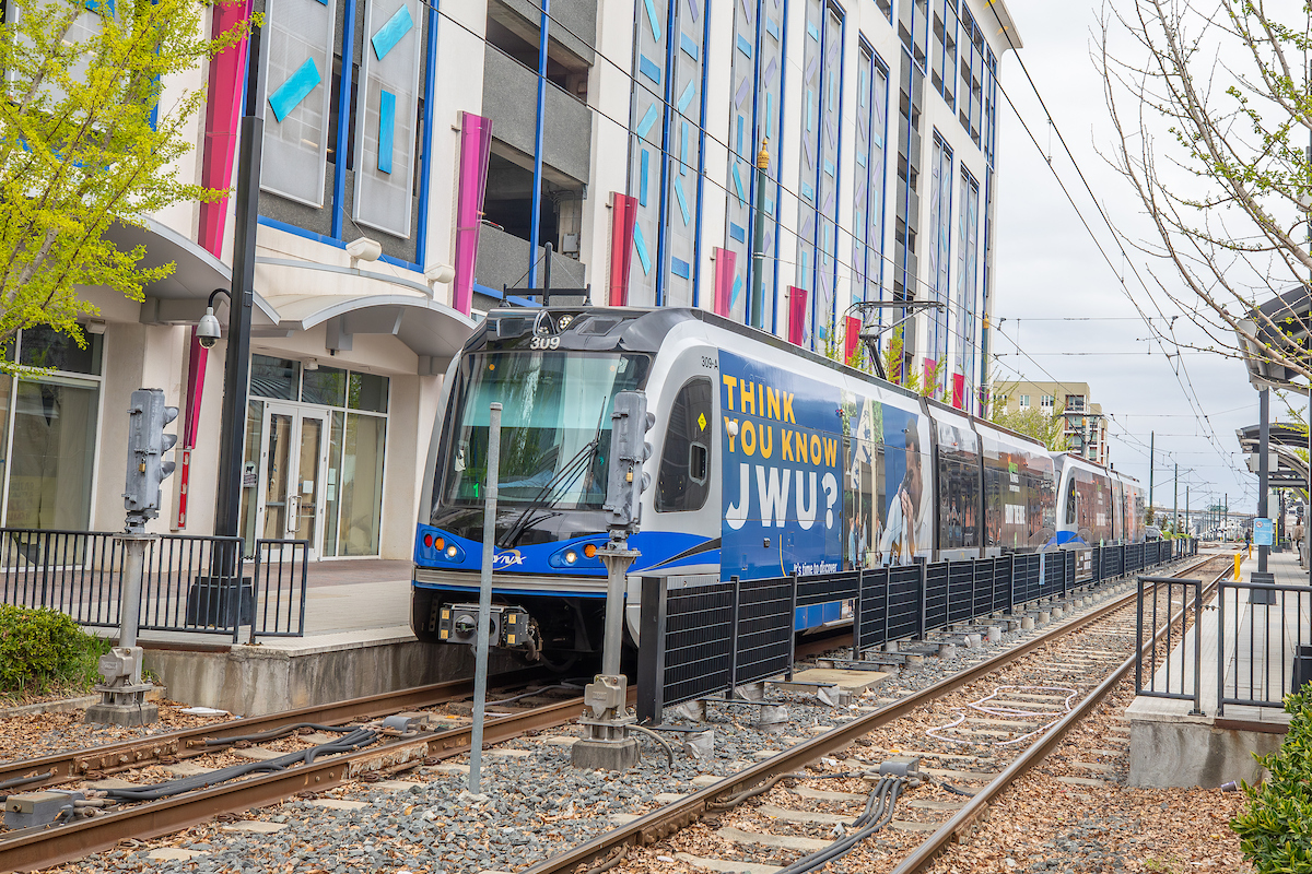 A modern light rail train with a "Think You Know JWU?" advertisement passes by a colorful building on an urban street.