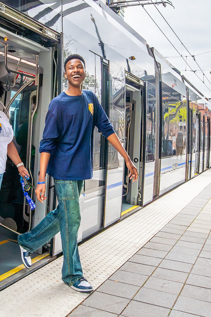 A person in a blue shirt and jeans steps off a modern tram at a station, smiling broadly.