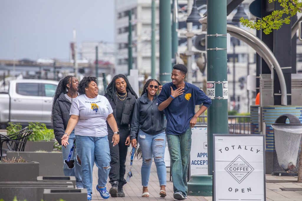 A group of five people walk together along a city sidewalk, smiling and chatting, with a coffee shop sign visible nearby.