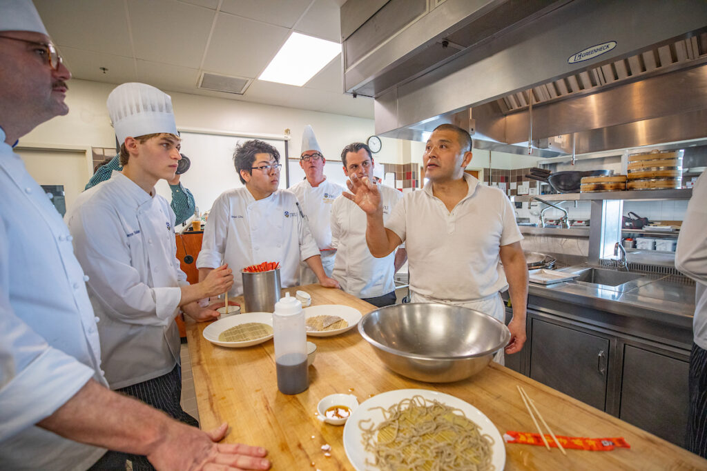 A chef instructs a group of trainees in a professional kitchen as they prepare noodles.