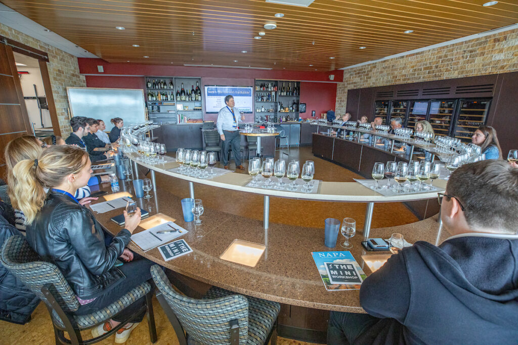 A group of attendees sits at a long curved table during a wine tasting workshop, with a presenter standing at the front near a screen and shelves of wine bottles.