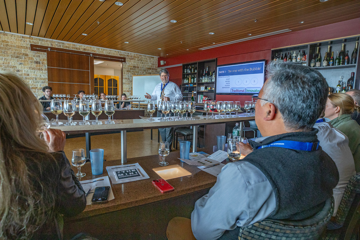 A group of attendees participates in a wine tasting session led by a presenter at a long table with numerous glasses of wine.