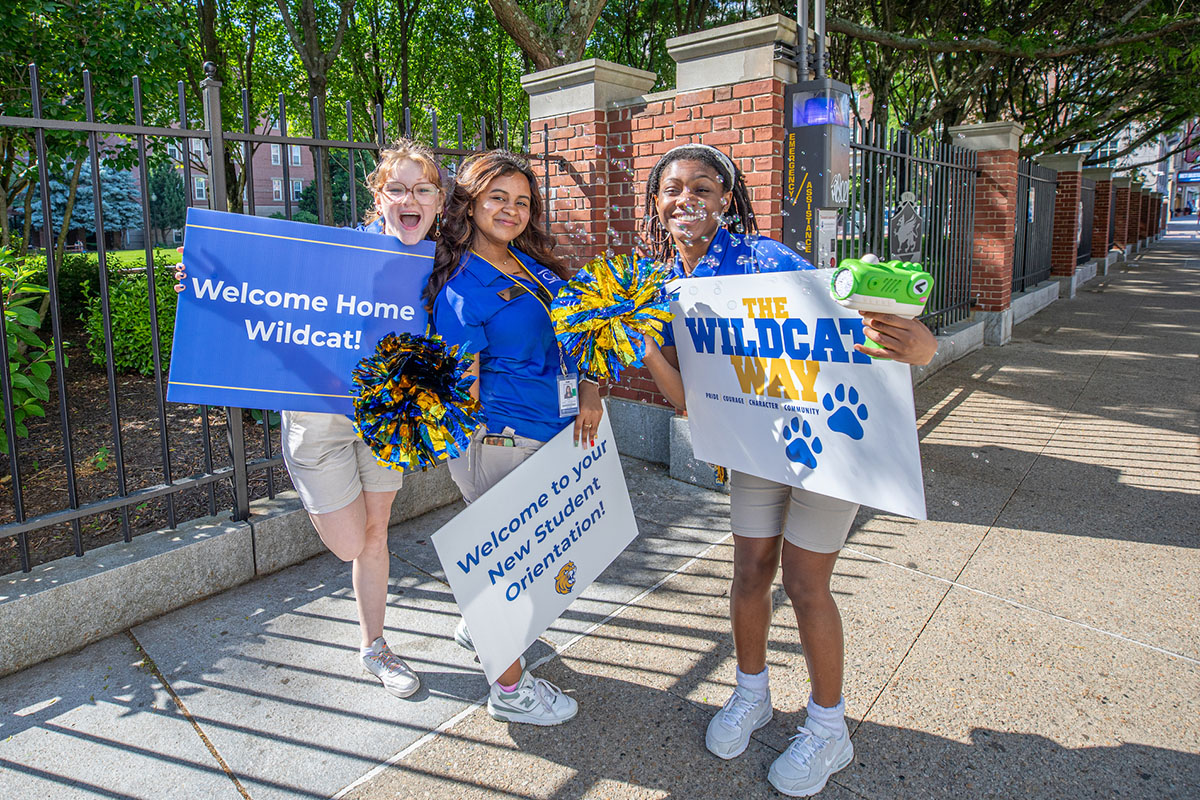 Three cat team members hold signs welcoming new students to campus, standing on a sunny sidewalk outside Gaebe Commons.