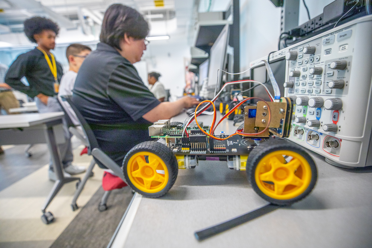 A person works on a small robot with yellow wheels in a lab setting, surrounded by electronic equipment and other individuals.