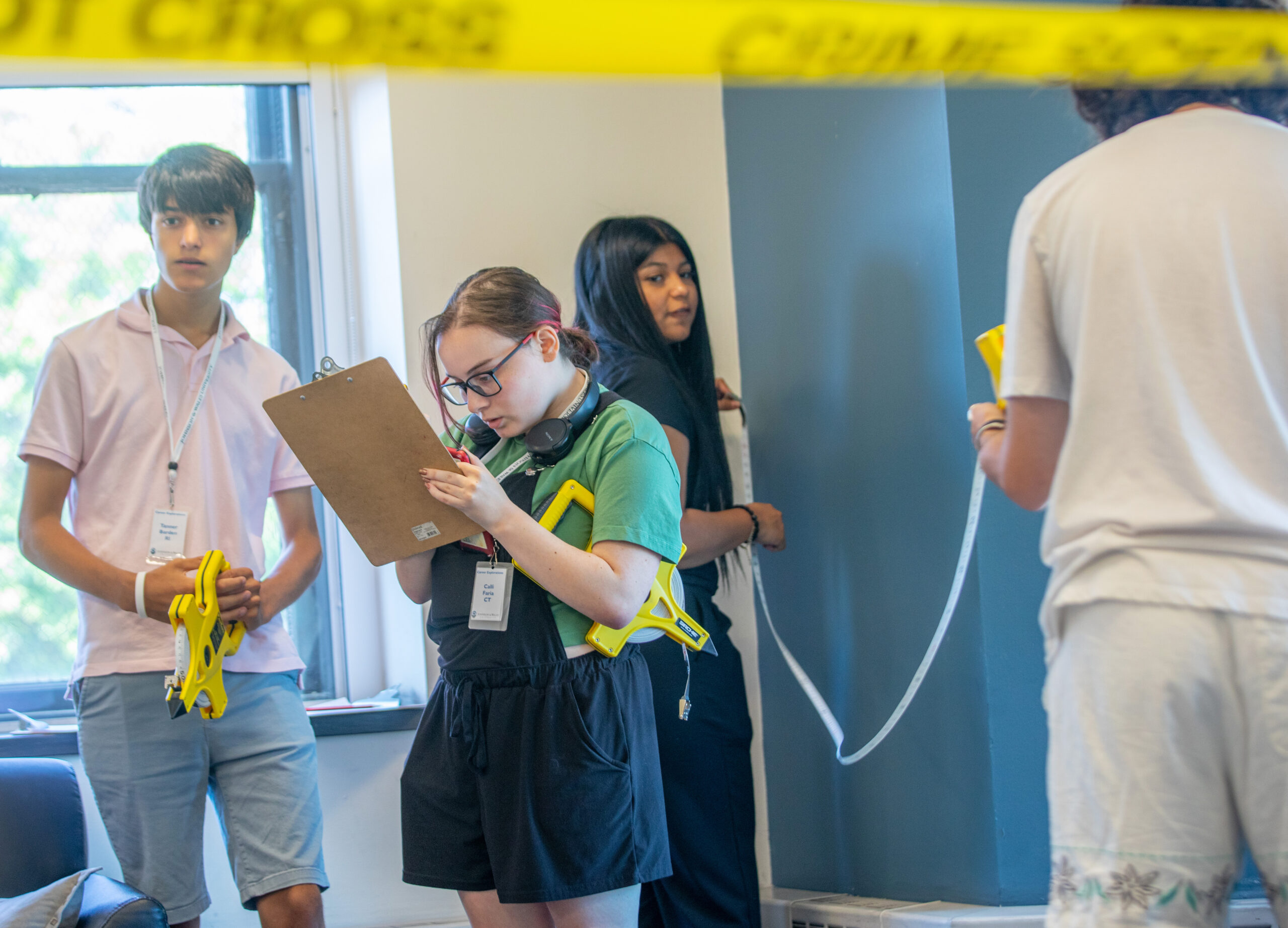 A group of young people are gathered inside a room with yellow crime scene tape, one person holding a clipboard while others observe.