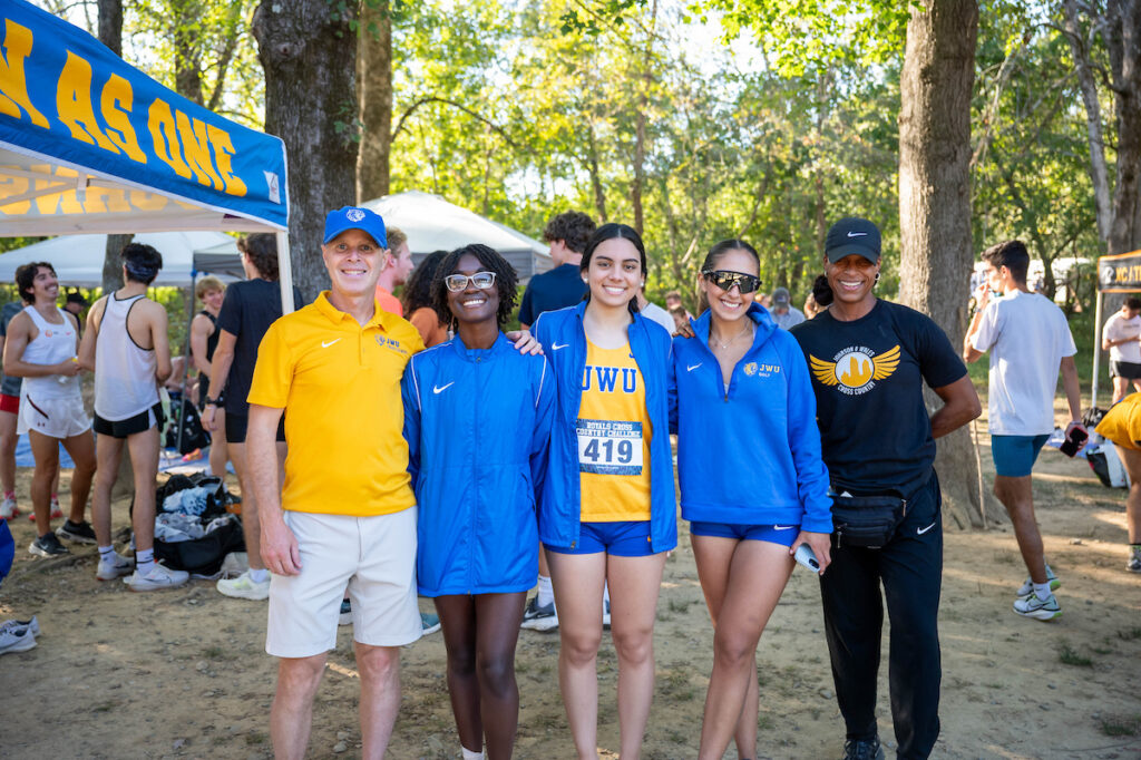 A group of four people, including athletes and staff, pose together at an outdoor cross-country event under a blue banner reading "RUN AS ONE."